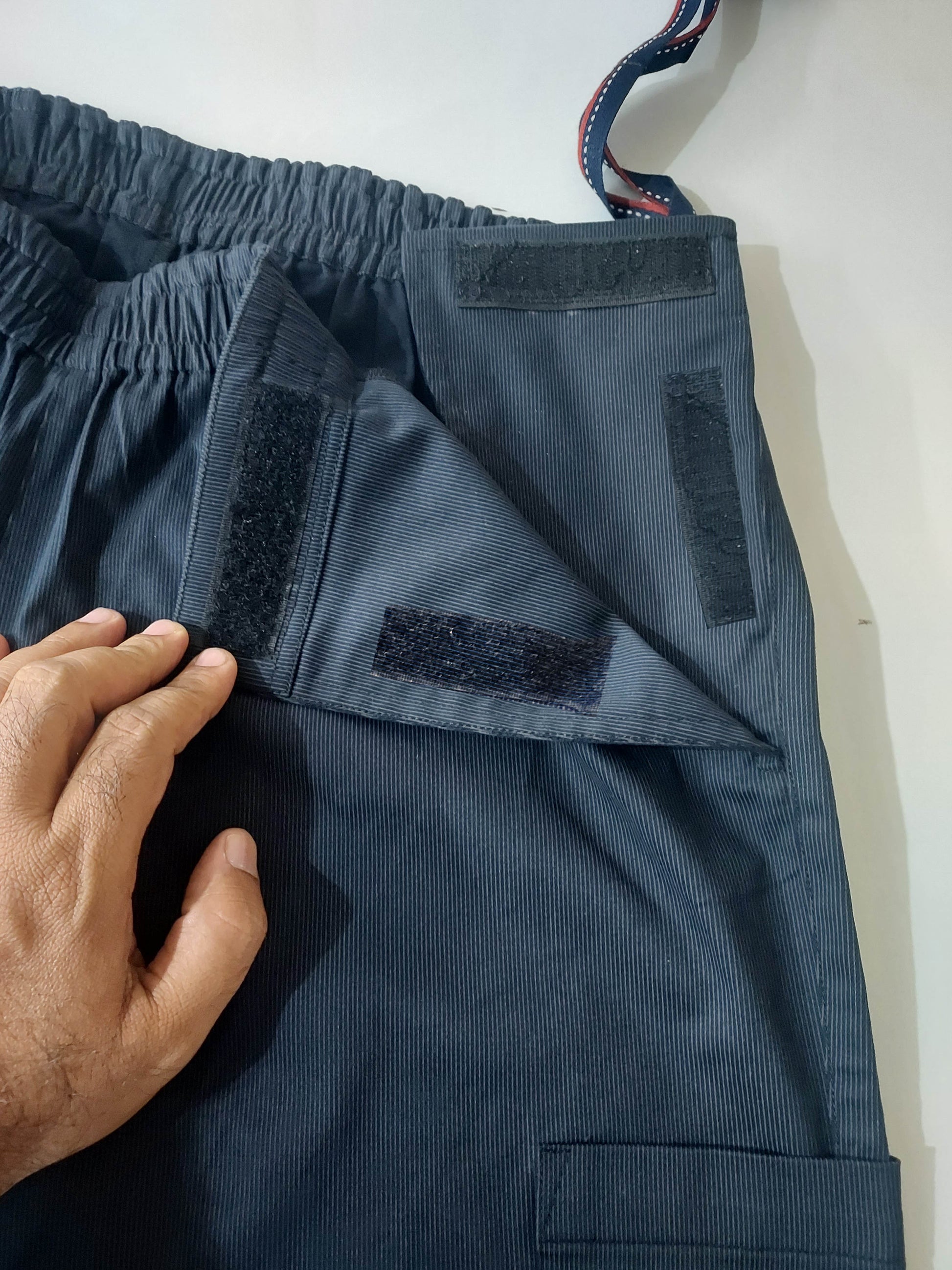 In this picture you can see the Wearer's left side around the waist and thigh region. there is an opening with visible velcros. one can see a hand opening the front. the pulling loops are visible. one can also see the some portion of the patch pocket. 